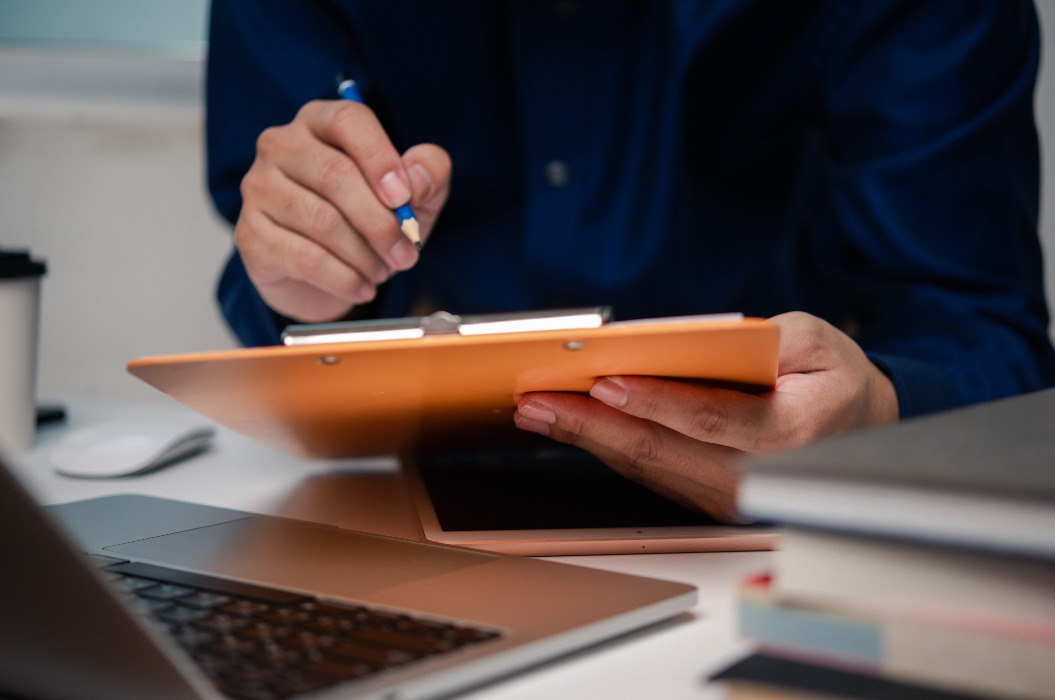 Ein Mann, der ein Klemmbrett mit einer Checkliste in der Hand hält; ein Mitarbeiter am Arbeitsplatz prüft Dokumente